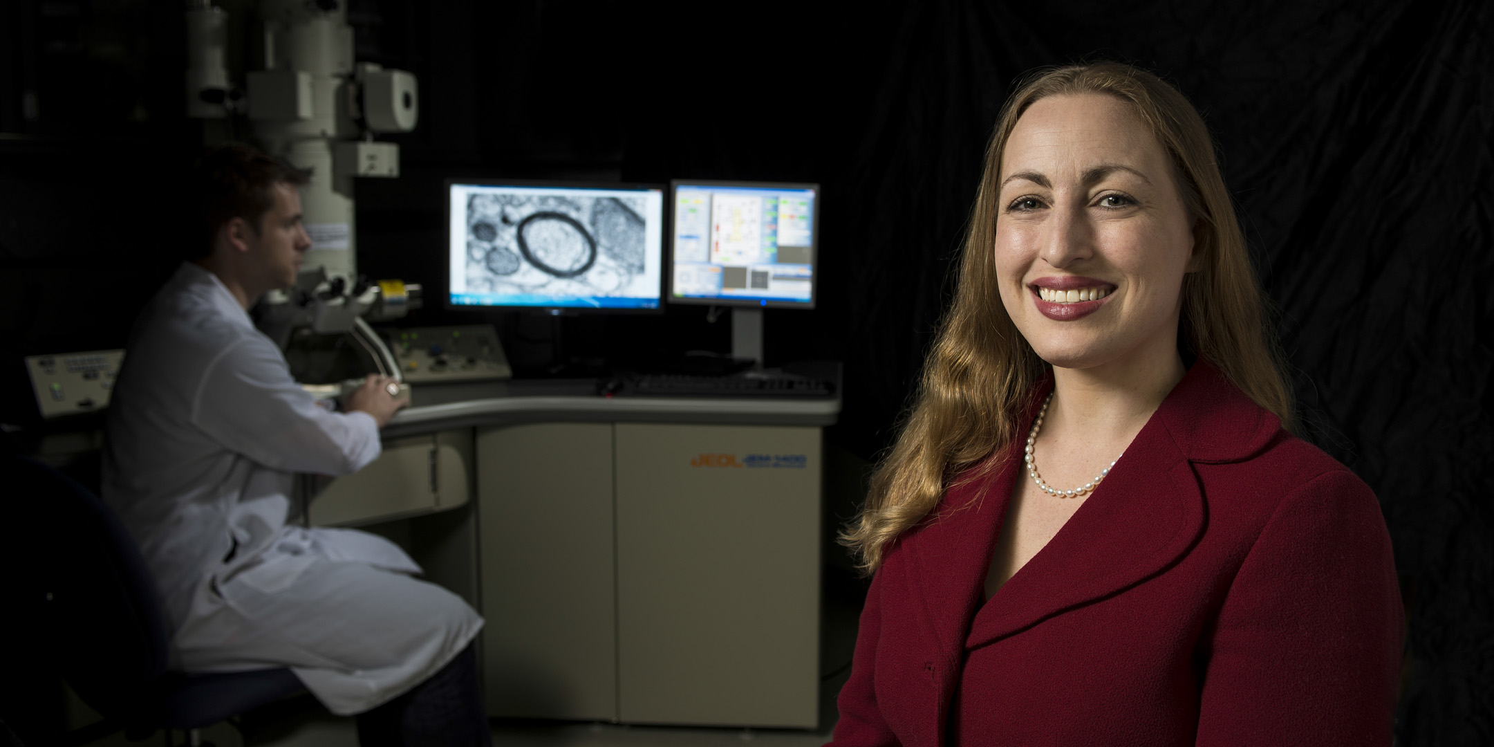 Michelle Monje stands in the foreground while a researcher examines an image of myelin in the background.