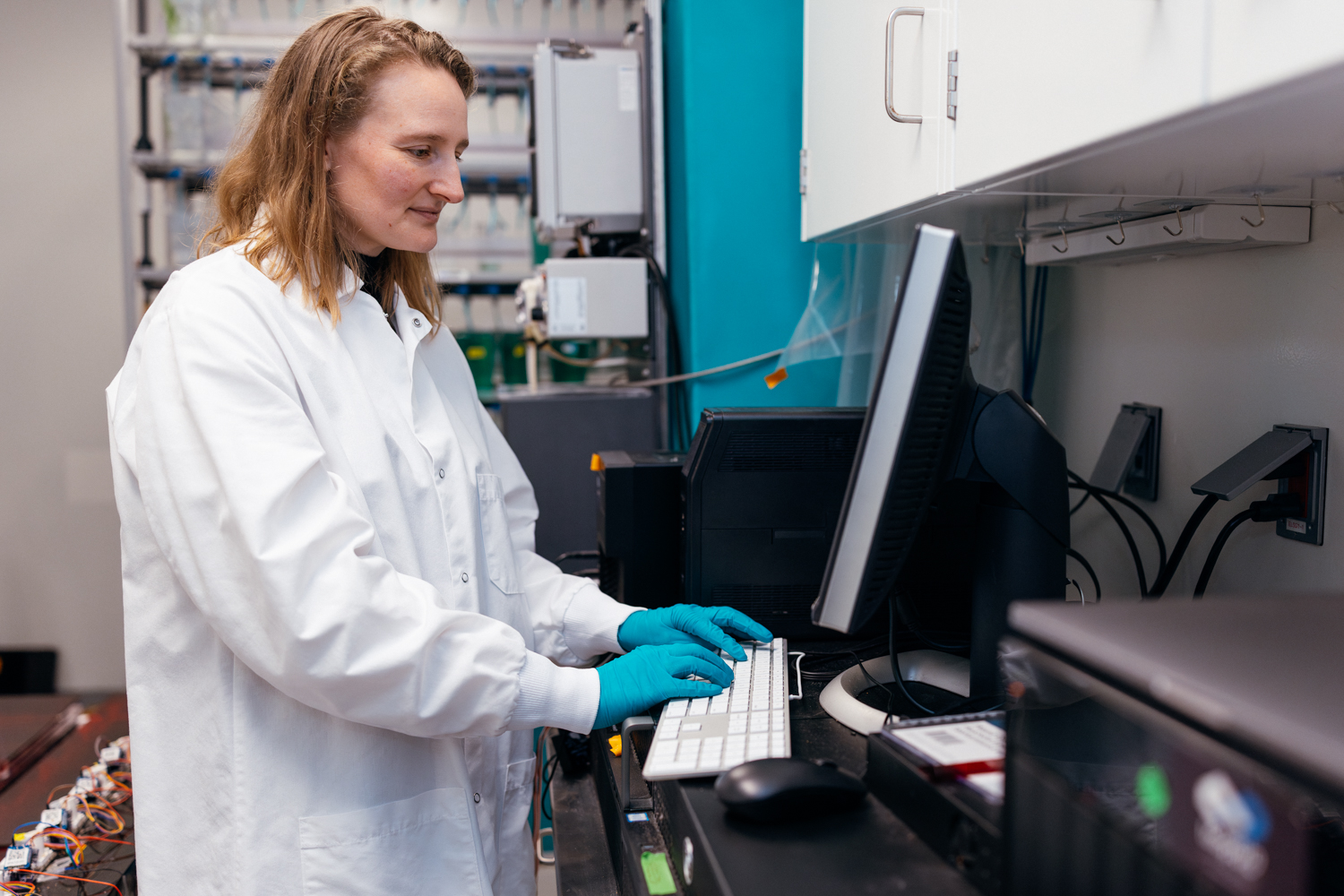 Claire Bedbrook working at a lab computer wearing a white lab coat and blue nitrile gloves.