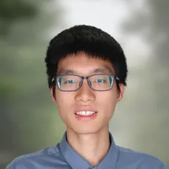 A professional headshot of Fei Deng, smiling warmly, wearing a blue button-up shirt. The background is softly blurred with green tones, giving a natural and professional appearance.