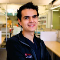 A professional headshot of Julian Garcia wearing a black jacket, standing with arms crossed in a lab setting.