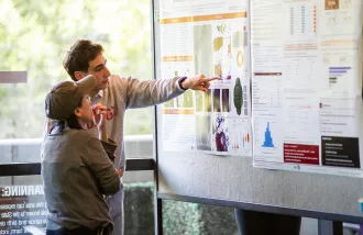 A researcher points to a poster describing his project while an onlooker listens.