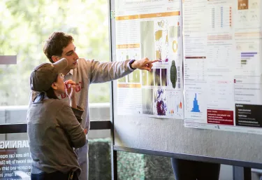 A researcher points to a poster describing his project while an onlooker listens.