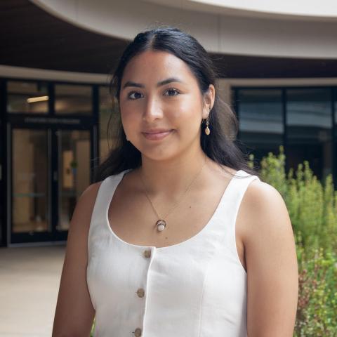 Wendy Paola Angeles stands outdoors near an entrance, wearing a white buttoned top and a necklace with a round pendant.