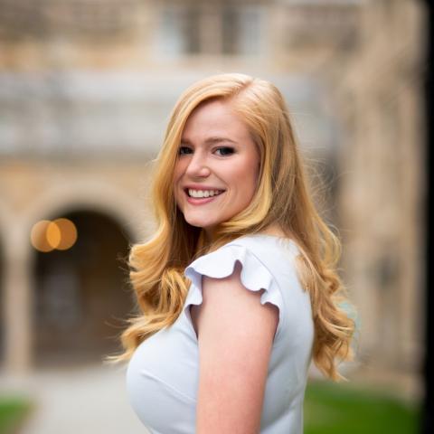 A professional headshot of Annie Goettemoeller, smiling and standing outdoors. She is wearing a light-colored blouse, and the background features blurred architecture and greenery for a natural and elegant feel.