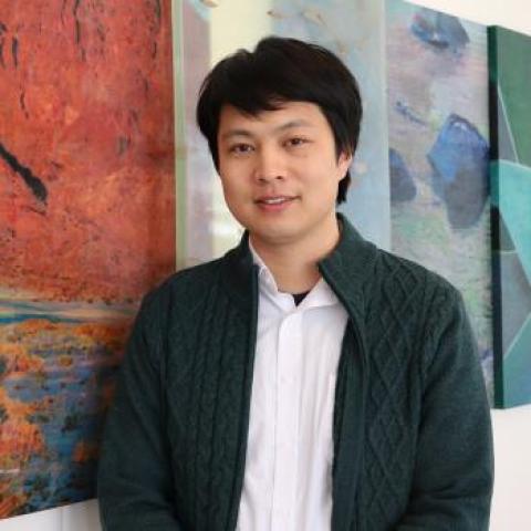 A professional headshot of Jian Xiong, wearing a dark green cardigan over a white shirt. He stands in front of colorful abstract artwork, offering a creative and formal atmosphere.