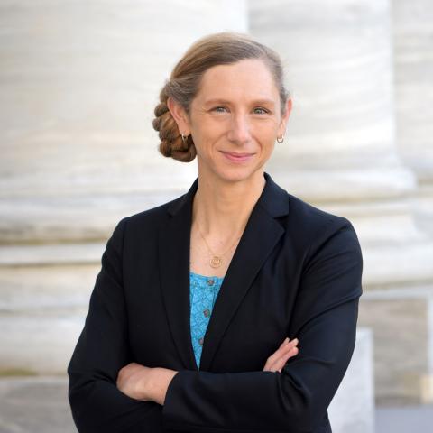 Picture of Dr. Ryann Fame. Dr. Fame is standing with her arms crossed and looking directly at the camera. She is smiling. She is wearing a black blazer and bright blue top underneath. 