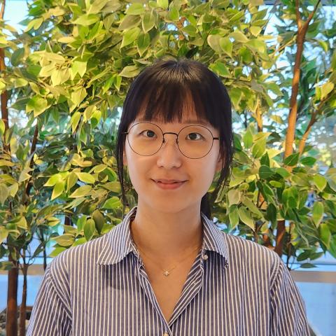 Ja Eun Choi wears glasses and a striped button down shirt and stands in front of green foliage while smiling at the camera