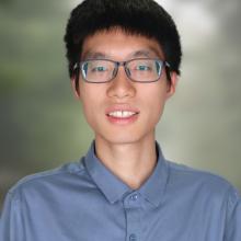 A professional headshot of Fei Deng, smiling warmly, wearing a blue button-up shirt. The background is softly blurred with green tones, giving a natural and professional appearance.