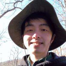 A candid outdoor photo of Hongfei Ji, smiling under a wide-brimmed green hat. The background features leafless trees against a bright blue sky, creating a natural and adventurous vibe.