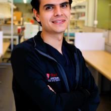 A professional headshot of Julian Garcia wearing a black jacket, standing with arms crossed in a lab setting.