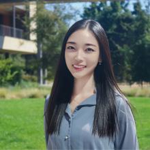 A professional headshot of Kelly Yoo, smiling outdoors with long straight black hair, wearing a light blue blouse. The background features a sunny green lawn and a building, creating a fresh and vibrant atmosphere.