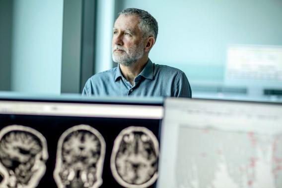 Michael Greicius stands behind computer screens displaying brain scans and text.
